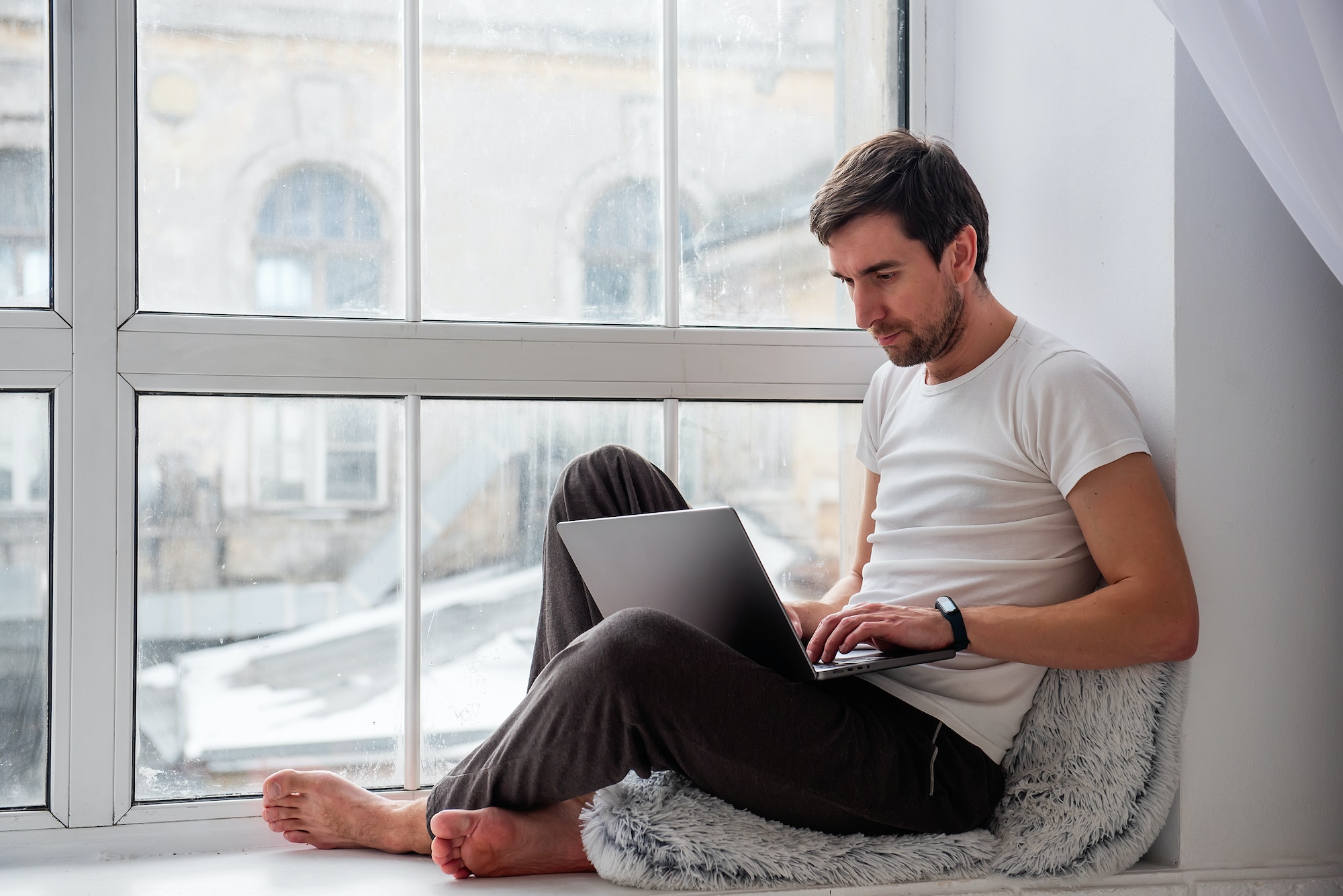 Young handsome man sits on wooden windowsill near the window in lotus position, works on gray laptop